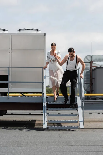 Elegante par de bailarines tomados de la mano mientras caminan por las escaleras en el techo - foto de stock