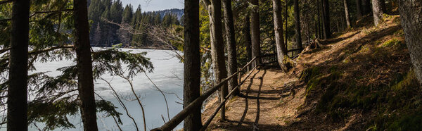 Walkway with sunlight near lake with ice in forest, banner 