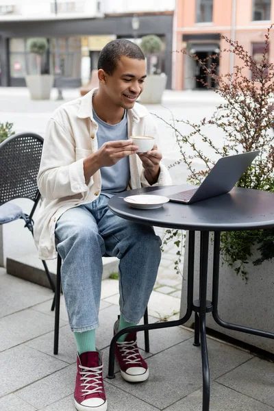 陽気なアフリカ系アメリカ人のフリーランサーがコーヒーを飲みながらカフェテラスでノートパソコンを使って — ストック写真