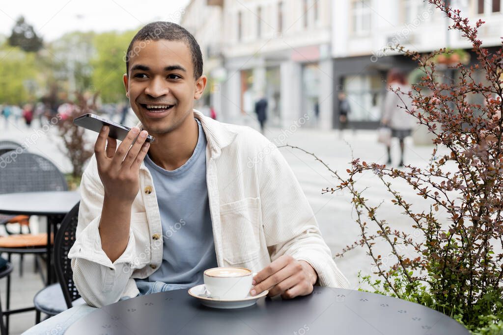 happy african american man recording voice message on smartphone near cup of coffee on table
