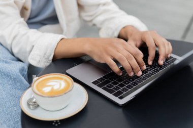 Kafe terasındaki cappuccino bardağının yanında dizüstü bilgisayarda çalışan Afrikalı Amerikalı serbest çalışanın...