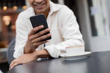Kesilmiş manzara gülümseyen Afrikalı Amerikalı adam kafenin terasındaki kapuçino bardağının yanında akıllı telefondan mesajlaşıyor. 