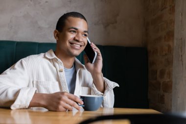 Gülümseyen ve Afro-Amerikalı genç bir adam akıllı telefondan konuşuyor ve kahve dükkanında bardak tutuyor. 