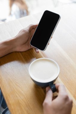Kapuçino bardağının yanında elinde boş ekranla akıllı telefon tutan bulanık Afro-Amerikan adam görüntüsü. 