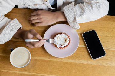 Afro-Amerikan bir adamın elinde çatal, yanında lezzetli bir tart ve boş ekranlı akıllı telefon tutuşu. 