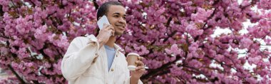 Gülümseyen Afrikalı Amerikalı adam elinde içecek ve akıllı telefonla konuşuyor. Çiçek açan kiraz ağacının yanında, pankartta.