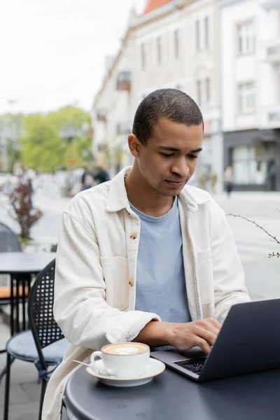 アフリカ系アメリカ人のフリーランサーがカフェテラスでカプチーノ近くのノートパソコンを使って — ストック写真