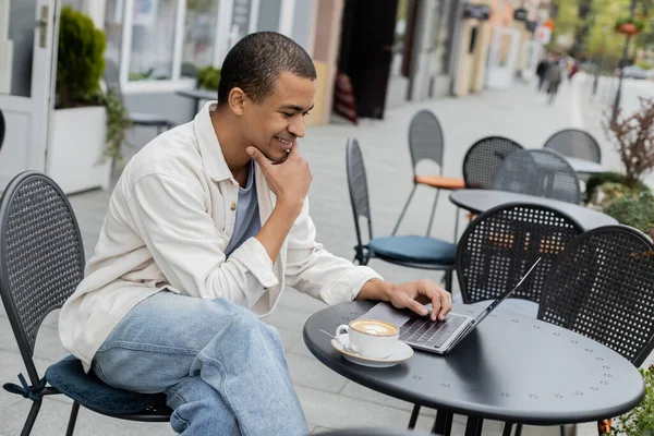 アフリカ系アメリカ人のフリーランスがカフェテラスでカプチーノのカップの近くのノートパソコンでリモートで働いています — ストック写真