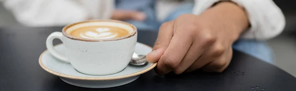 stock image cropped view of african american man holding saucer with cup of cappuccino on summer terrace, banner