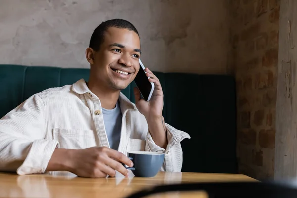 Sonriente Joven Hombre Afroamericano Hablando Teléfono Inteligente Sosteniendo Taza Cafetería — Foto de Stock