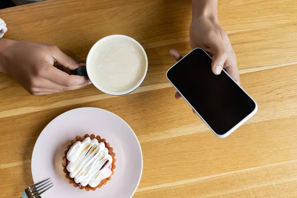 アフリカ系アメリカ人男性がスマートフォンを持っていてカプチーノが美味しいタルトの近くにあり — ストック写真