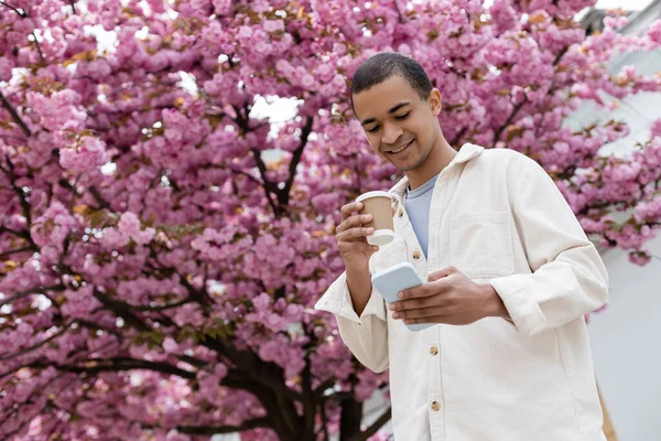在粉红樱桃树附近拿着咖啡去用智能手机的快乐的非洲裔美国人的低视角照片 — 图库照片