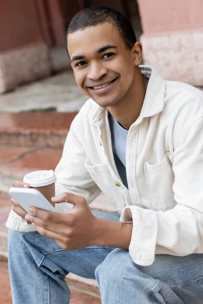 Feliz Homem Americano Africano Segurando Bebida Takeaway Smartphone Enquanto Sentado — Fotografia de Stock