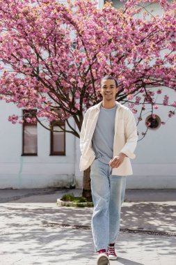 Gülümseyen Afro-Amerikan gömlekli adam. Cebinde Japon kiraz ağacının yanında yürüyor.