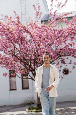 Tişörtlü neşeli Afro-Amerikan adam, cebinde Japon kiraz ağacının yanında yürüyor.