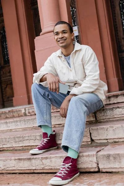 Pełna Długość Uśmiechnięty Afroamerykanin Człowiek Koszuli Kurtka Trzymając Smartfon Siedząc — Zdjęcie stockowe