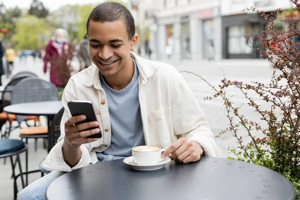 Feliz Homem Americano Africano Mensagens Texto Smartphone Perto Cappuccino Mesa — Fotografia de Stock