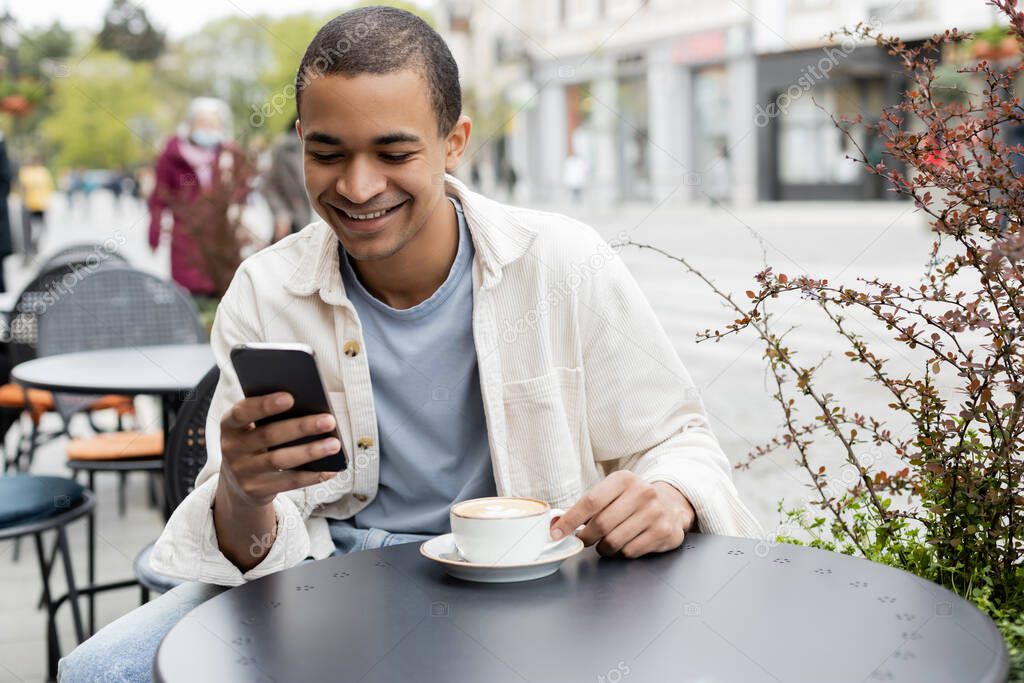 happy african american man texting on smartphone near cappuccino on table
