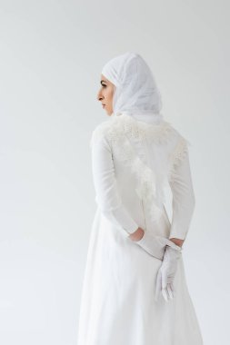 Eldivenli ve beyaz elbiseli Müslüman gelin elleri arkada, gri renkte.