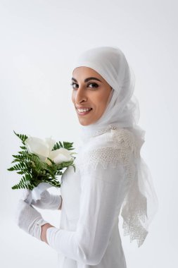 Eldivenli ve gelinlikli neşeli Müslüman gelin Gri renkte zambak çiçekleri tutuyor