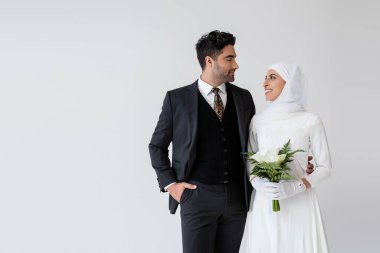 Müslüman gelin gelinlik içinde elinde kalla zambağı buketi ve gri renkte izole edilmiş takım elbiseli mutlu damada bakıyor.