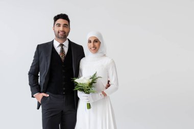 Eldivenli ve gelinlikli Müslüman gelin elinde bir buket zambak, gri elbiseli mutlu damat.