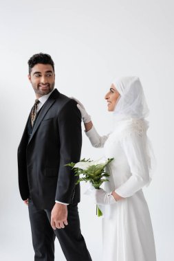 Gelinlik içinde mutlu Müslüman gelin elinde bir buket zambak ve gri elbiseyle damadın arkasında duruyor.
