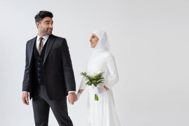 Gülümseyen Müslüman gelinlik içinde bir buket zambak ile damat ile el ele tutuşan gri elbiseli gelin.