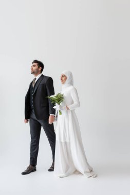 Gülümseyen Müslüman gelinlik içinde bir buket zambak ile gri üzerinde mutlu damadın elini tutuyor.
