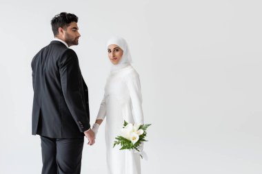Gülümseyen Müslüman gelinlik içinde bir buket zambak ile Gri üzerinde izole damat ile el ele.
