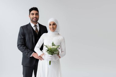 Müslüman damat, gri renkte izole edilmiş bir buketle gelinlik giymiş neşeli geline sarılıyor. 