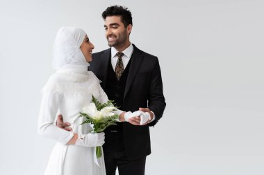 Müslüman damat, gri renkte izole edilmiş bir buketle gelinlik içinde gülümseyen gelinin elini tutuyor. 