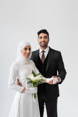 Müslüman damat beyaz elbiseli mutlu gelinin elini tutuyor ve gri renkte buketi var. 