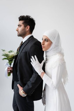 Müslüman gelin tesettür içinde damada sarılıyor ve buketi gri renkte izole ediyor. 