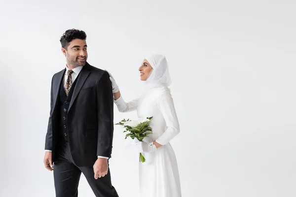 結婚式のドレスで笑顔のイスラム教徒の花嫁は カラユリの花束を保持し 灰色で隔離されたスーツで新郎の後ろに立って — ストック写真