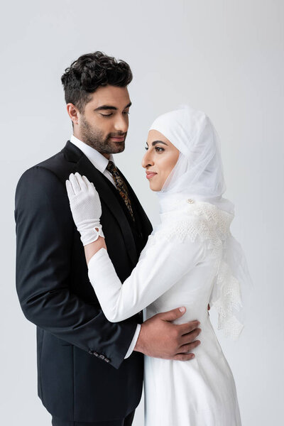 young muslim bride in hijab and wedding dress and groom hugging isolated on grey