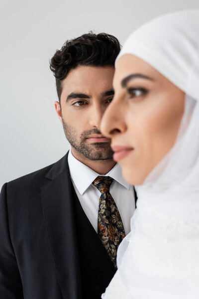 Muslim man looking at blurred bride on foreground isolated on grey