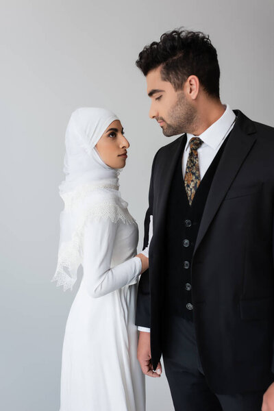 woman in wedding dress and hijab standing near husband in suit isolated on grey 