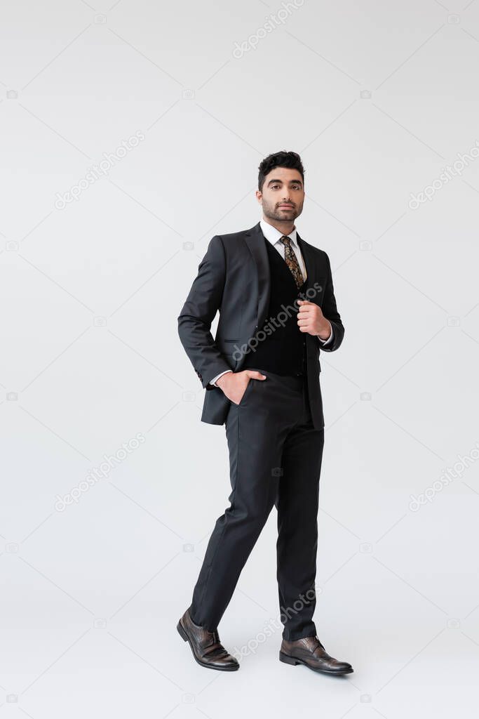 full length of muslim groom in formal wear standing with hand in pocket on grey 