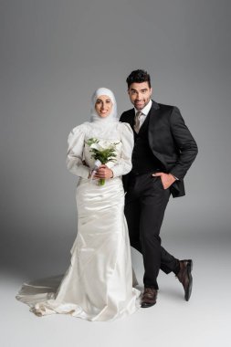 Takım elbiseli mutlu damat, gri buketli Müslüman gelinin yanında poz veriyor.