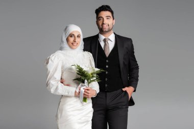 Takım elbiseli Müslüman damat, gri renkte izole edilmiş bir düğün buketiyle mutlu gelin pozu veriyor.