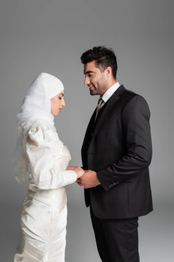 Müslüman gelin gelinlik içinde ve takım elbiseli neşeli damat gri renkte el ele tutuşmuş.