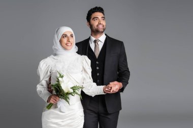 Mutlu damat rüya gibi Müslüman gelinin elini tutuyor. Gri üzerine saklanmış zambak buketi ile.