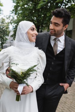 Mutlu damat cepte poz veriyor ve düğün buketiyle tesettürlü Müslüman geline bakıyor. 