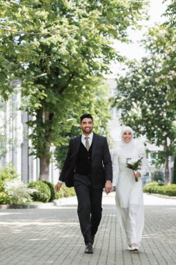 Mutlu damat gülümseyen Müslüman gelinle el ele tutuşuyor ve düğün buketi ile birlikte yürüyor.