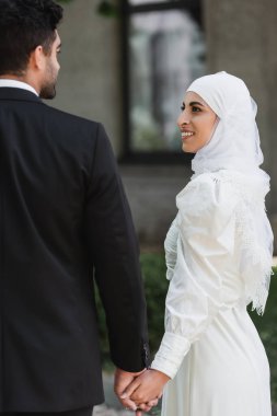Damat, dışarıda gelinlik giymiş, gülümseyen Müslüman gelin ile el ele tutuşuyor.  