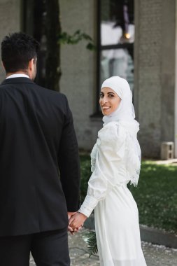Damat, gelinlik giymiş, neşeli Müslüman gelinle el ele tutuşuyor ve birlikte yürüyor. 