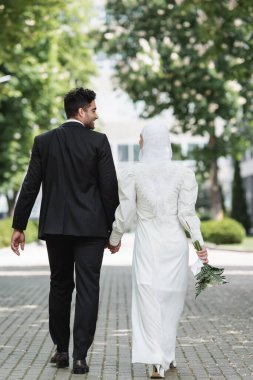 Mutlu damat, düğün buketli Müslüman kadınla el ele tutuşuyor ve birlikte yürüyor.