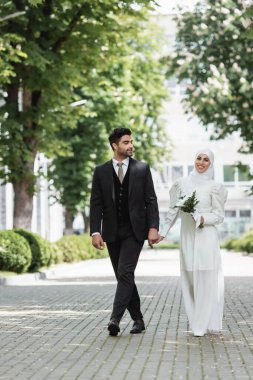 Mutlu damat gülümseyen Müslüman gelin ile el ele tutuşuyor düğün buketi ile tesettüre 