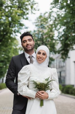 Gelin tesettürlü neşeli Müslüman ve beyaz elbiseli, damadın yanında buket tutuyor. 